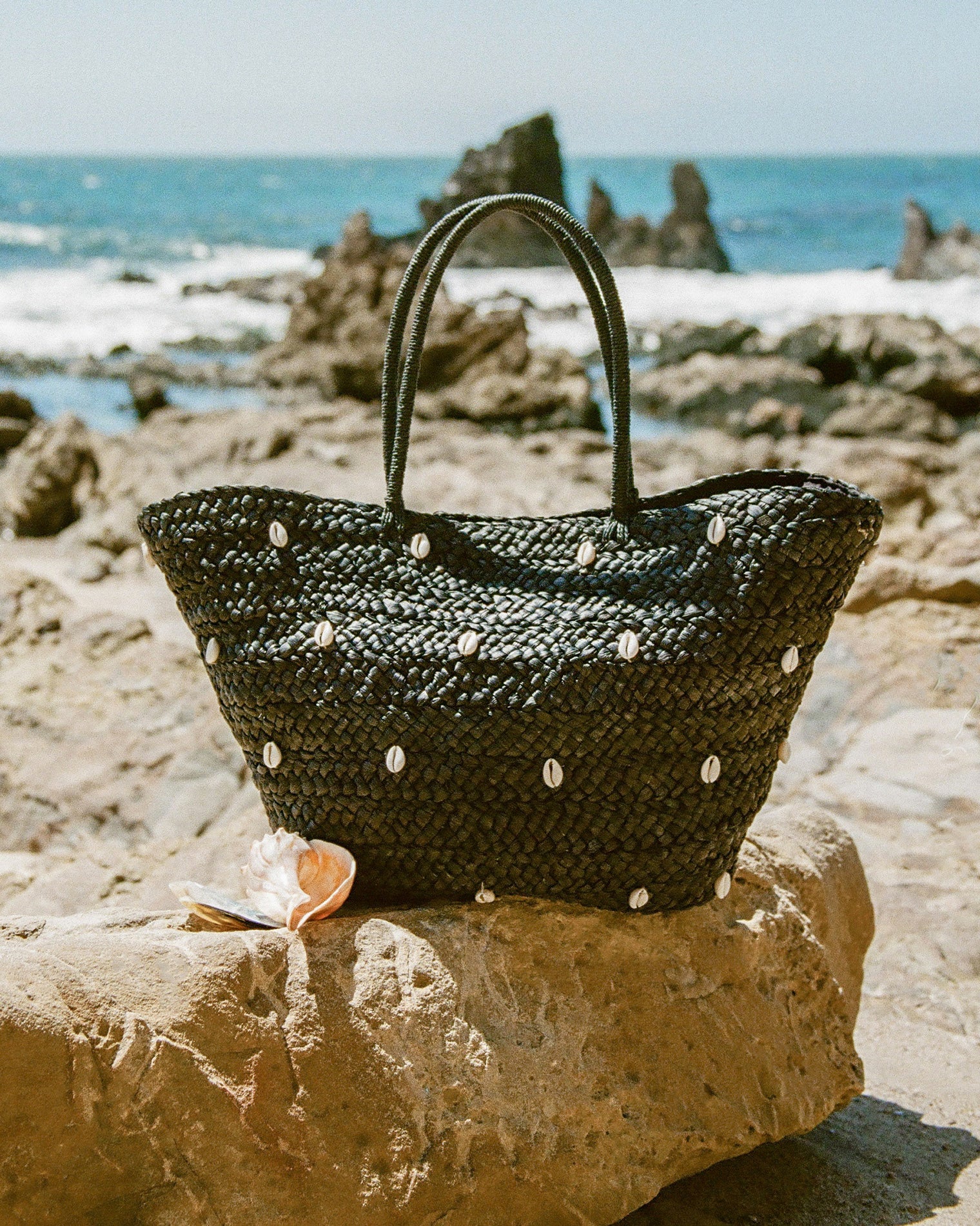 Baltic Seas Tote - Black Lifestyle image of the Baltic Seas Tote in black resting on a rocky coastal ledge with a seashell beside it and the ocean in the background.