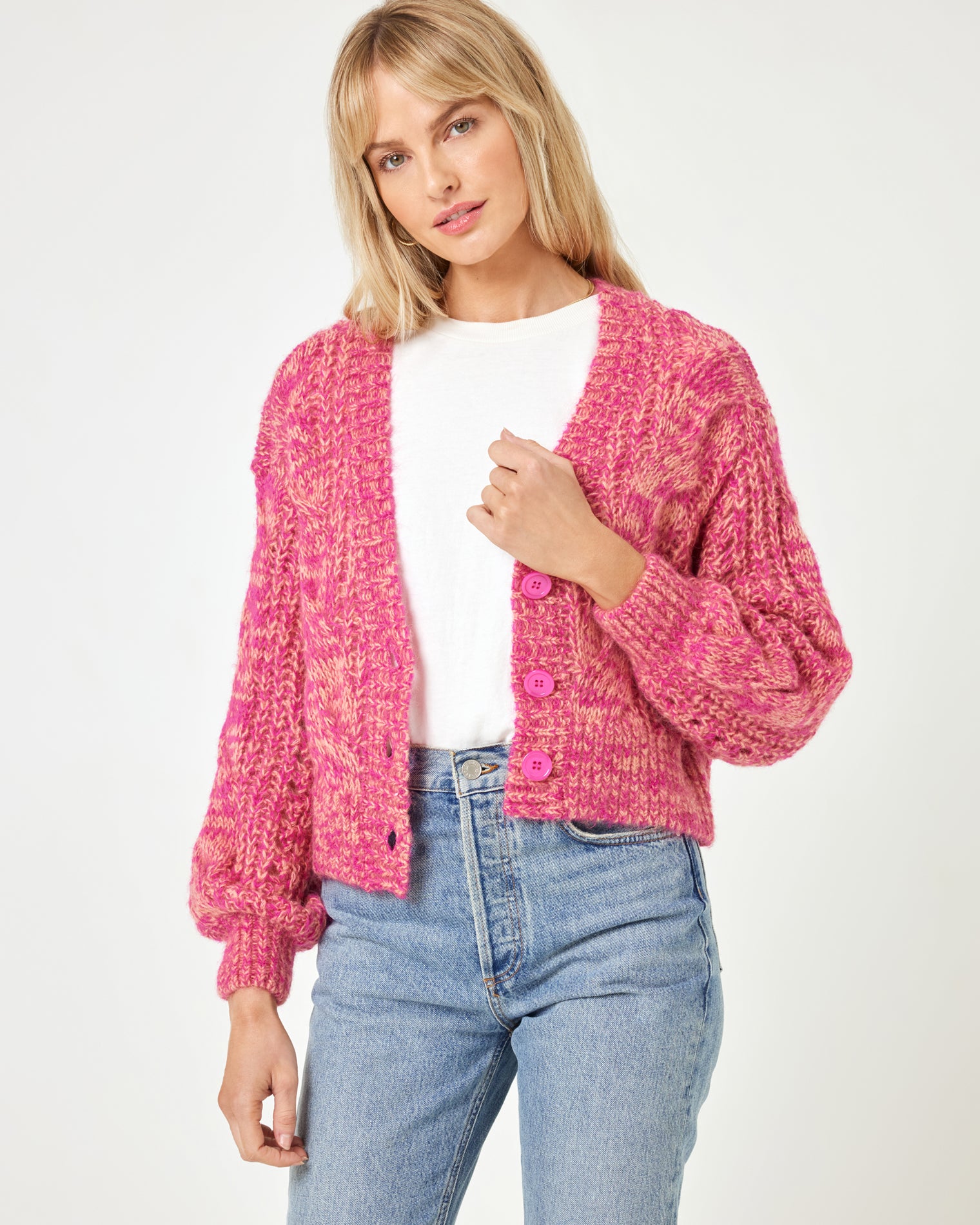Strawberry Fields Cardigan - Strawberry Marl Front view of a model wearing the LSPACE Strawberry Fields Cardigan in strawberry marl, a pink cable-knit button-front V-neck cardigan with pink buttons, styled open over a white tee and jeans with sneakers on a white background.