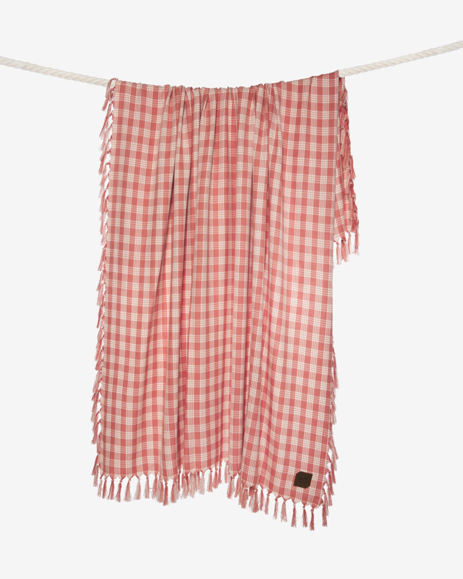 Slowtide Palaka Lightweight Beach Blanket - Guava The Slowtide Palaka Lightweight Beach Blanket in guava hanging on a rope, showing the pink gingham check pattern with fringe.