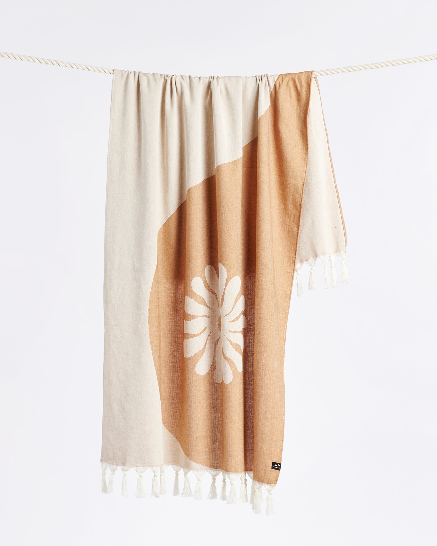 Slowtide Botanic Balance Turkish Towel - Brush The Slowtide Botanic Balance Turkish Towel in brush hanging on a rope, showing the yin-yang botanical pattern with daisy motif in tan and cream with fringe.