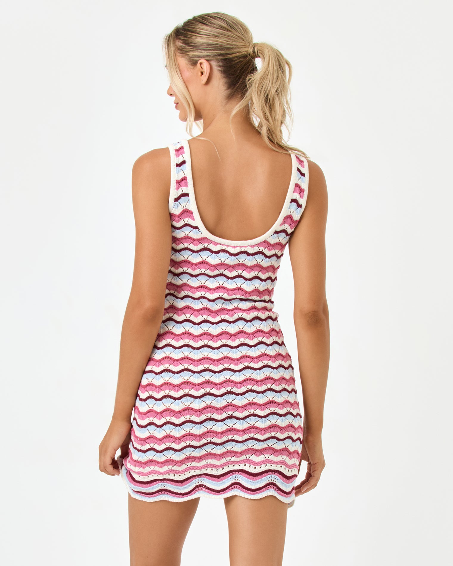 Sunchaser Dress - Sand In Hand Back view of a model wearing the LSPACE sand in hand Sunchaser Dress, a crochet knit mini dress showing the scoop back neckline and multicolor wavy stripe pattern in pink, navy, and white, on a neutral studio background.