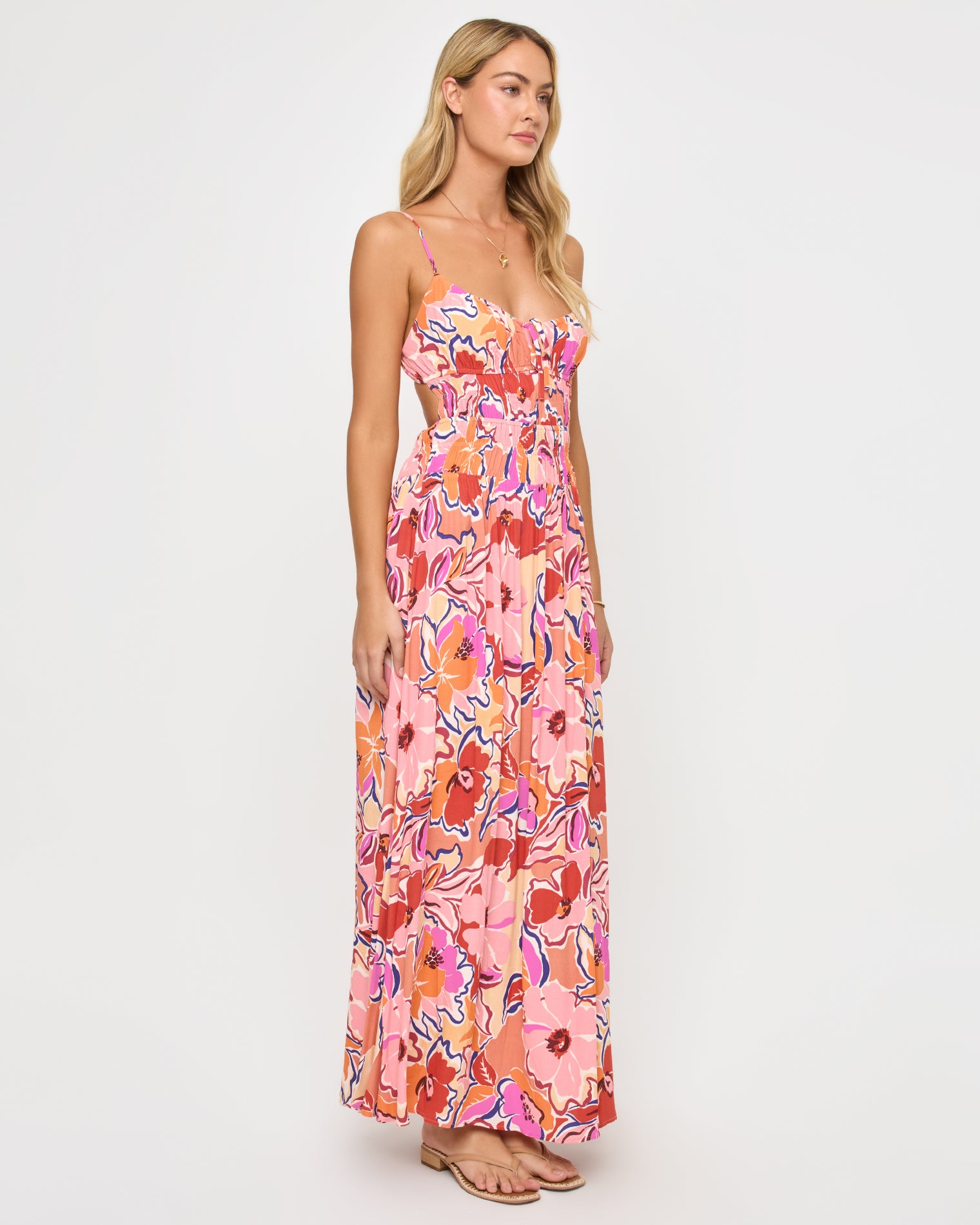Tala Dress - Lost In The Blooms Angled view of a model wearing the LSPACE Tala Dress in lost in the blooms, showing the side open-back cutout of the retro floral maxi dress with the vibrant pink, orange, and red bloom print.