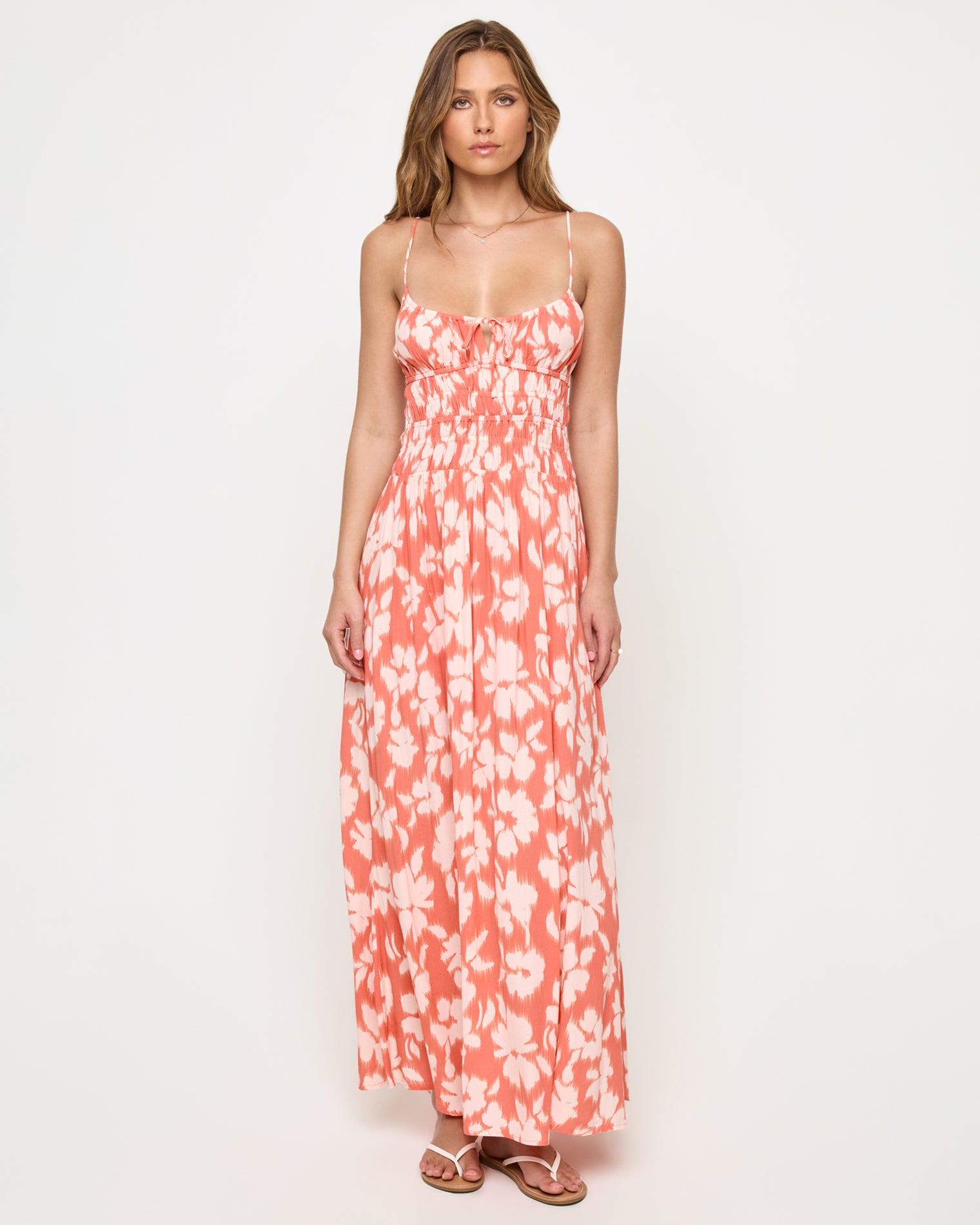 Tala Dress - Vine Vignette Front view of a model wearing the LSPACE Tala Dress in vine vignette, a spaghetti-strap maxi dress with a front tie detail, smocked empire waist, and coral and white floral print.