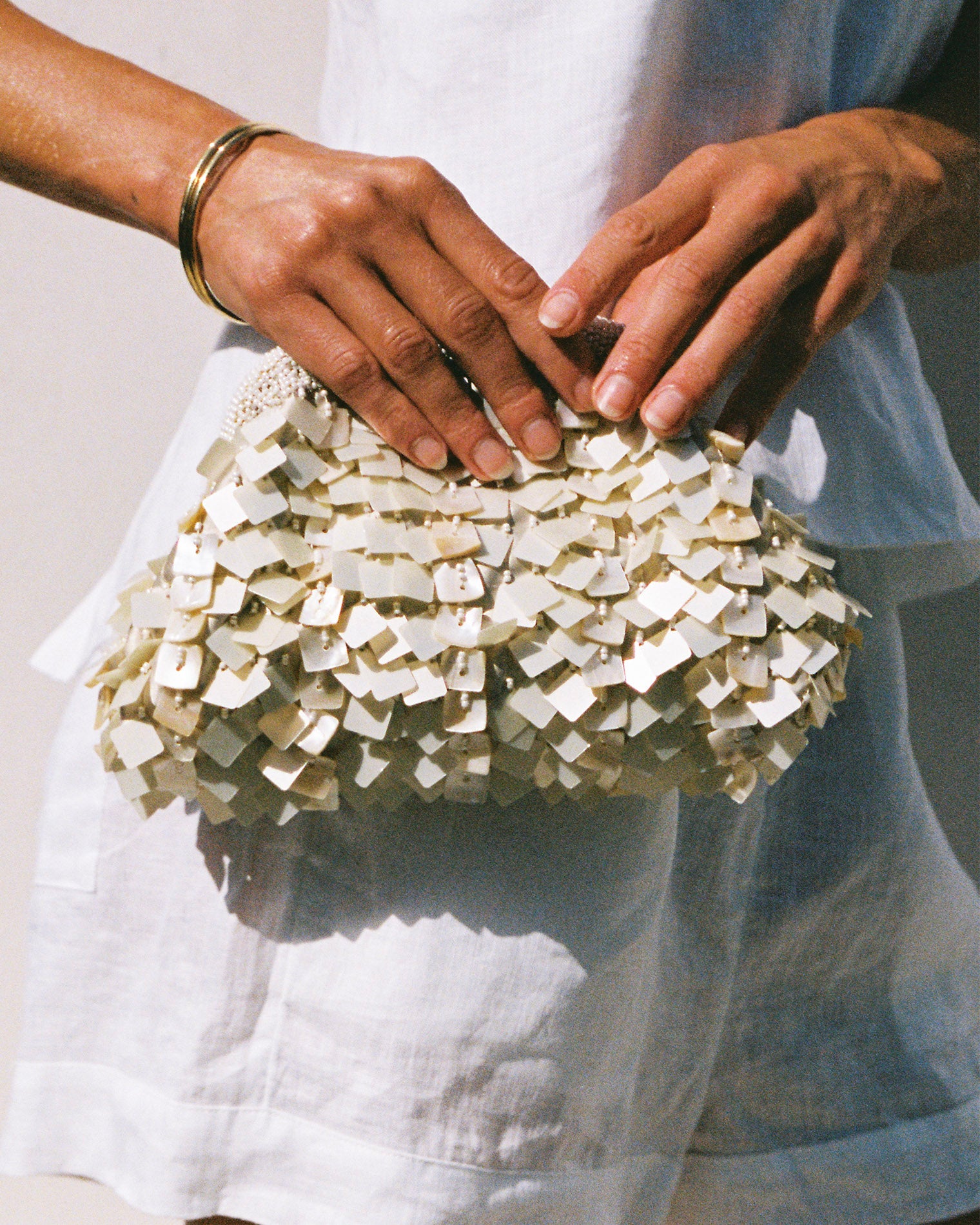 Vienna Clutch - Cream Model holding the Vienna Clutch in cream against a white linen outfit, showing the layered shell paillette texture.