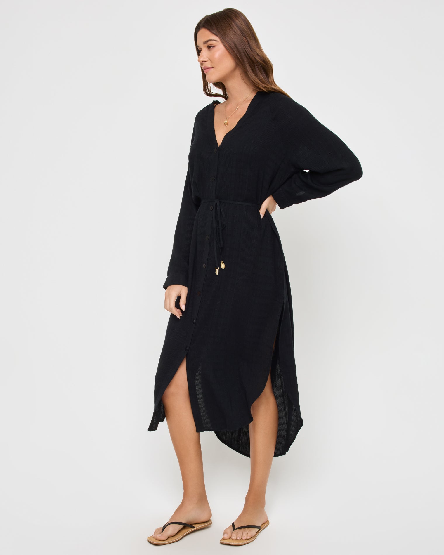 Yoli Shirtdress - Black Side view of a model wearing the LSPACE Yoli Shirtdress in black, showing the side profile with the V-neckline, long dolman sleeves, drawstring waist, and high-low hem with a side slit.