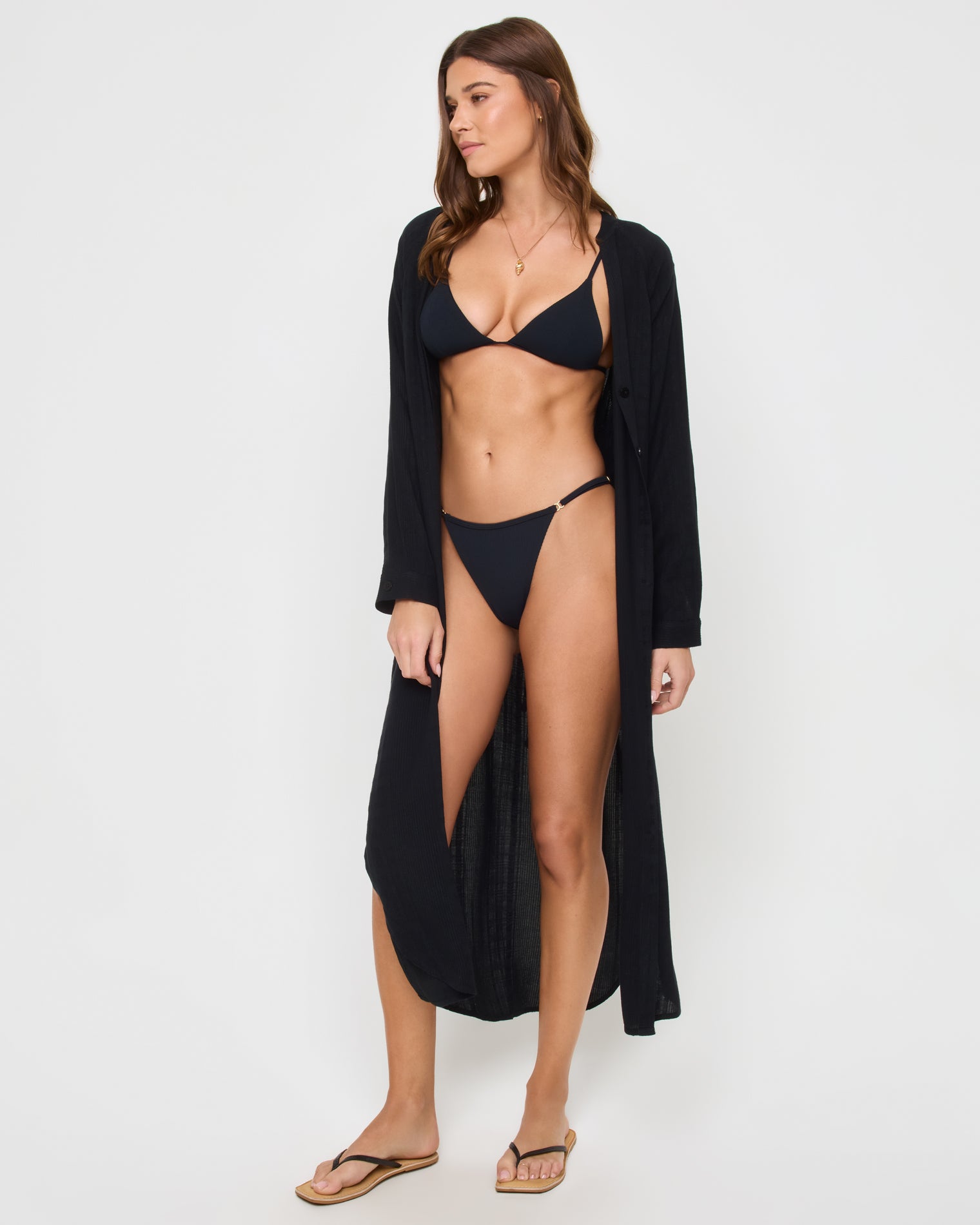 Yoli Shirtdress - Black Angled front view of a model wearing the LSPACE Yoli Shirtdress in black, styled fully open as a maxi-length beach coverup over a black bikini, showing the long flowing silhouette.