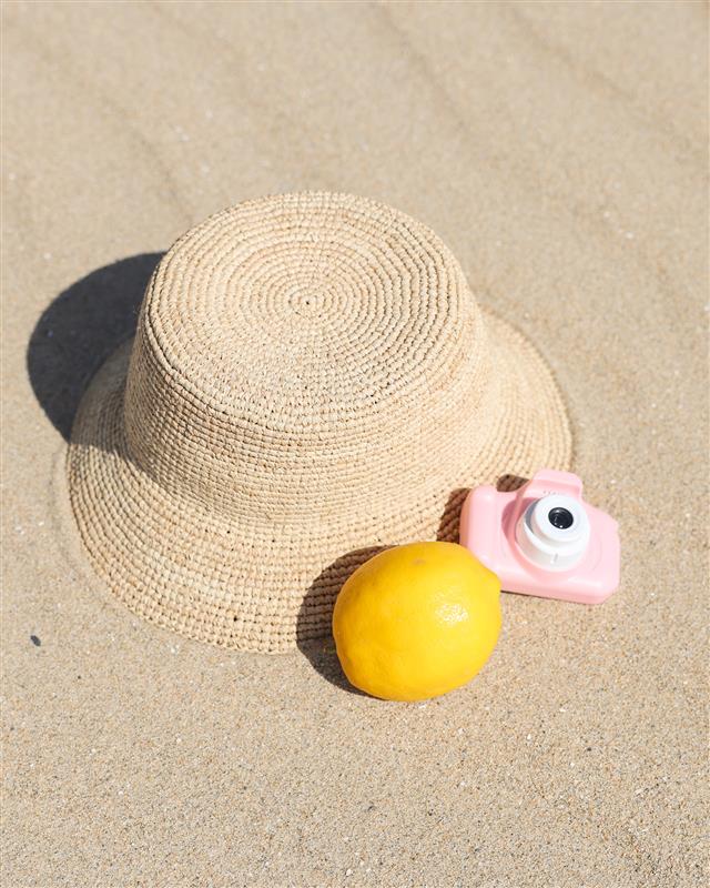 Isadora Mini Bucket Hat - Natural LSPACE Isadora mini bucket hat in natural, off-model flat lay with lemon and toy camera on sand.